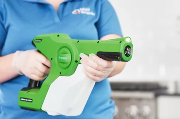 Cleaning professional holding a green electrostatic disinfectant sprayer in a kitchen setting.